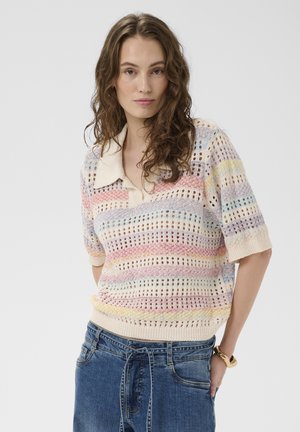 Kvinde med krøllet hår iført en pastelfarvet, stribet, åben strikket kortærmet sweater og blå denimjeans, stående med hænderne bag ryggen.