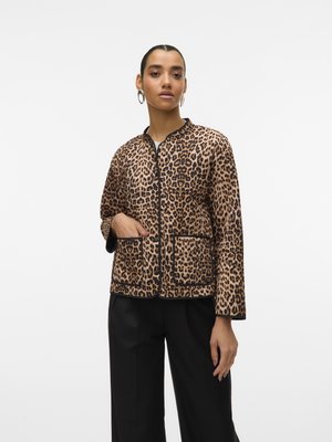 Femme portant une veste en imprimé léopard avec des bordures et des poches noires, associée à un pantalon noir, se tenant contre un fond blanc uni.