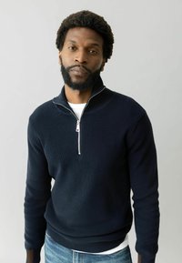 Dunkelblauer, gerippter Strick-Halbzipperpullover mit einem silbernen Reißverschluss, getragen über einem weißen Hemd, kombiniert mit hellblauen Jeans.