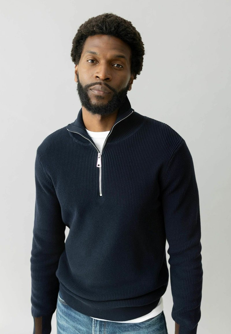Dunkelblauer, gerippter Strick-Halbzipperpullover mit einem silbernen Reißverschluss, getragen über einem weißen Hemd, kombiniert mit hellblauen Jeans.