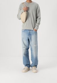 Persoon in een lichtgrijze trui, lichtblauwe versleten jeans, beige sneakers en met een beige schoudertas over één schouder.