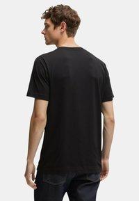 Giovane uomo con capelli corti castani che indossa una semplice maglietta nera a maniche corte, voltato di spalle su uno sfondo chiaro e uniforme.