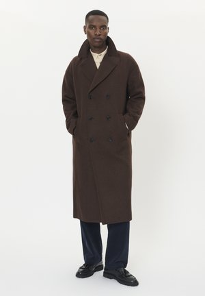 Brown wool double-breasted coat with a wide collar, featuring black buttons and side pockets, worn over dark pants and black shoes.