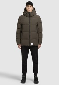 Puffer jacket in olive green with a hood, featuring a zip closure and side pockets. Paired with black sweatpants and sneakers.