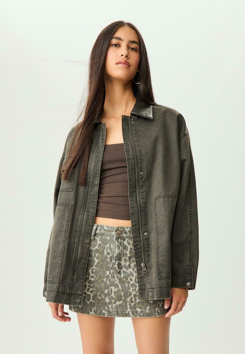 Giacca di jeans nera oversize con chiusura a bottoni e tasche applicate, abbinata a un top corto marrone e una gonna grigia con stampa leopardata.