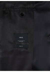 Intérieur en tissu noir d'une veste avec deux étiquettes cousues, dont l'une porte la mention "MNG" et "Slim Fit", et un bouton noir au-dessus des étiquettes.