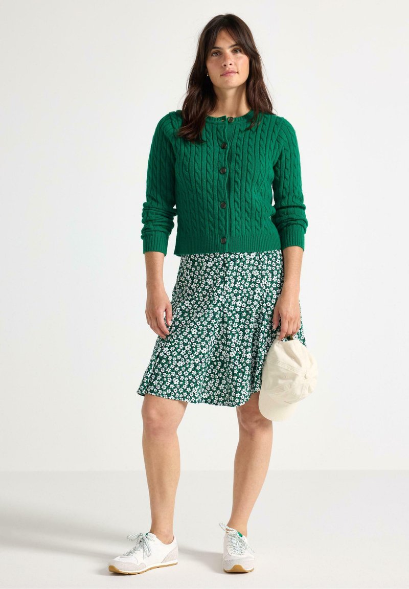 Grøn kabelstrik cardigan med brune knapper, parret med en blomstermønstret nederdel. Hvide sneakers og en beige kasket holdt i hånden.