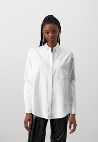White button-up shirt made of lightweight fabric, featuring a single chest pocket and long sleeves for a relaxed fit.