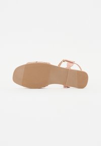 Platt sandal med gummisula, ljusrosa remmar och ett litet spänne, visad från botten på en vit bakgrund.