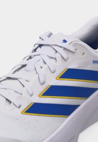 Λευκό αθλητικό παπούτσι με μπλε και κίτρινη τριπλή ρίγα, λευκά κορδόνια και υφασμάτινο επάνω μέρος με υφή, που εμφανίζει μερική πλευρική και κορυφαία όψη.