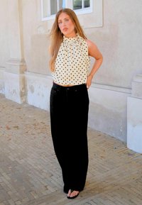Γυναίκα στέκεται έξω κοντά σε έναν τοίχο, φοράει λευκό αμάνικο τοπ με πουά, μακριά μαύρη φούστα και μαύρα σανδάλια, με μακριά μαλλιά κάτω.