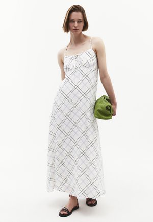 Mujer con un vestido largo blanco con cuadros negros, sosteniendo un bolso de mano verde con textura, usando sandalias negras, de pie frente a un fondo blanco.