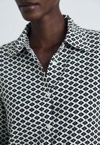 Person wearing black and white patterned collared shirt with small floral shapes and buttoned front, visible from the neck to mid-torso.