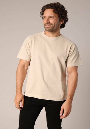 Beige katoenen T-shirt met korte mouwen en een ronde hals, in combinatie met zwarte broek, gepresenteerd op een mannelijke model tegen een neutrale achtergrond.