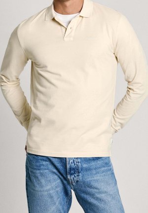 Mężczyzna w kremowej koszulce polo z długim rękawem i niebieskich dżinsach, z rękami opartymi na biodrach na jednolitym tle.