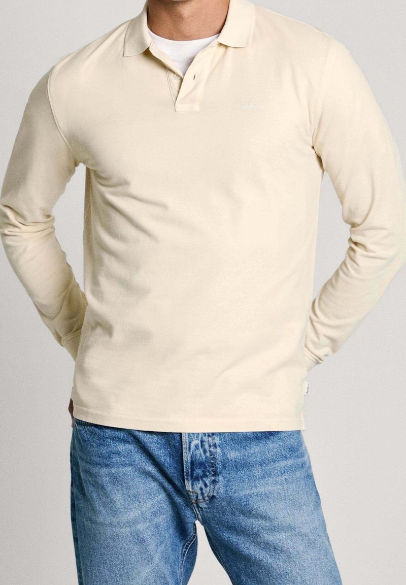 Homme portant un polo crème à manches longues et un jean bleu, les mains posées sur les hanches, sur un fond uni.