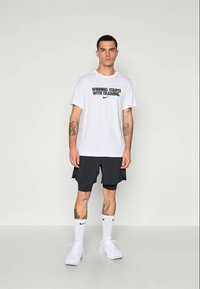 Mann iført en hvit "Winning Starts With Training" T-skjorte, svarte lagdelte shorts, hvite Nike-sokker og hvite treningssko, som står mot en ensfarget vegg.
