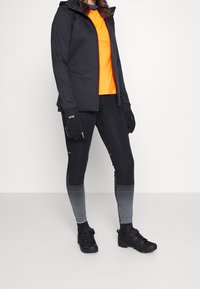 Svart jacka med huva över en orange tröja, i kombination med leggings i svart och grått gradient mönster och svarta skor. Tillbehören inkluderar svarta handskar.