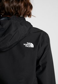Schwarze wasserdichte Jacke mit Kapuze, auf der Rückseite mit einem auffälligen weißen Logo. Glatte Textur, minimalistisches Design und sichtbare Nahtdetails.
