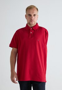 Rød polo-shirt med krage, kortærmede, og tre knapper. Har et lille logo på venstre side. Glat bomuldsstruktur.