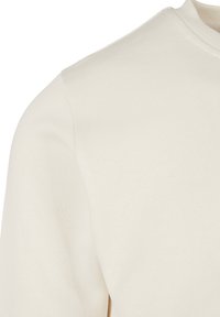 Gros plan sur un sweat-shirt crème à col rond, montrant l'épaule, la manche et une partie du col côtelé.