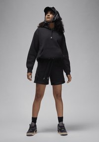 Hoodie preto com um bolso frontal, combinado com calções pretos. O modelo usa sapatos pretos com uma sola clara e um boné preto com um lenço estampado.