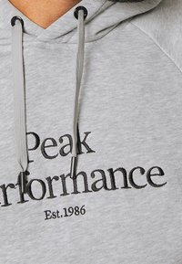 Hoodie gris clair avec cordons, présentant un texte brodé noir "Peak Performance Est. 1986" sur la poitrine.