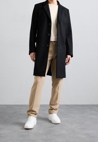 Abrigo de lana negro con un corte entallado, dos botones y bolsillos laterales, combinado con pantalones beige y zapatillas blancas sobre un fondo gris.