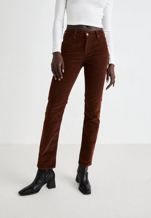 Femme portant un pantalon en velours côtelé marron, des bottines noires à talons et un top court blanc à manches longues sur un fond blanc uni.