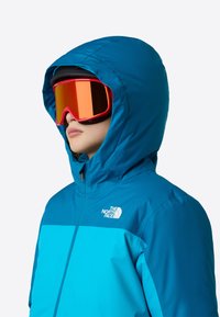 Jachetă izolată albastră cu glugă, cu un design de panouri contrastante. Asortată cu ochelari mari de schi cu lentile roșii. Branding subtil pe jachetă.