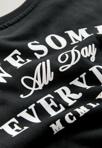 Čierne tričko z textilu s bielym textom, na ktorom je fráza "ÚŽASNÉ CELÝ DEŇ KAŽDÝ DEŇ." Text je zobrazený v rôznych typoch písma a veľkostiach.