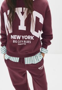 Felpa bordeaux con il bold bianco "NYC" e il testo "NEW YORK"; sovrapposta a una camicia a righe verdi e bianche sui polsini. Joggers coordinati sotto.