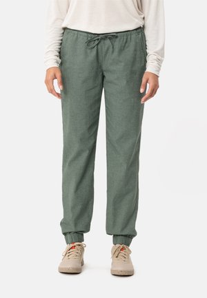 Persoon draagt groene broek met trekkoord en elastische boorden en beige sneakers, staand tegen een effen achtergrond.