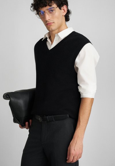 Czarny dzianinowy kamizelka z dekoltem w kształcie litery V, noszona na białej koszuli. Osoba trzyma teksturowany czarny worek i ma na sobie czarne spodnie.