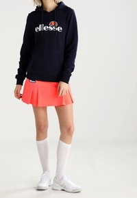 Sudadera náutica con un logo blanco, combinada con una falda plisada en color coral brillante. Medias blancas hasta la rodilla y zapatillas deportivas blancas completan el atuendo.
