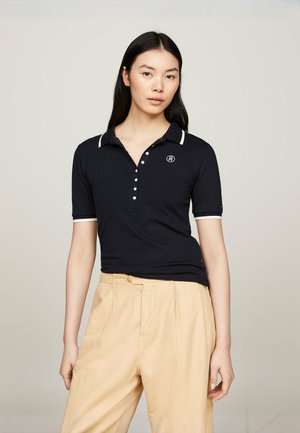 Polo shirt blu navy con colletto e dettagli bianchi sulle maniche, dotato di una pattina con bottoni e logo sul petto sinistro. Abbinato a pantaloni beige.