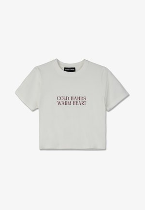 Camiseta blanca cortada con mangas cortas que presenta texto en color burdeos que dice "MANOS FRÍAS, CORAZÓN CALIENTE" en una fuente serif en negrita.