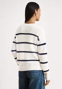 Pletene svetre v krémovej farbe s námorníckymi vodorovnými pruhmi, vybavené rebrovaným golierom a manžetami a pohodlným strihom. Nosené s modrými džínsami.