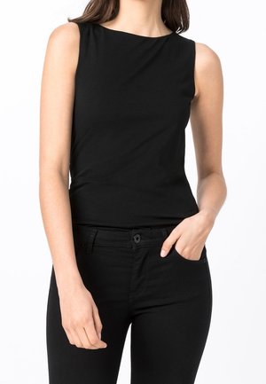 Femme portant un haut noir sans manches et un pantalon noir, une main dans la poche, sur un fond blanc uni.