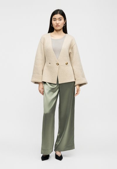 Γυναίκα που φοράει μπεζ μάλλινο σακάκι με κουμπιά, ελαφρύ ριμπ τοπ, παντελόνι σατέν ελιάς με πλατιά πόδια και μαύρα μυτερά παπούτσια μπροστά σε λευκό φόντο.