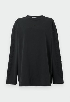 Vrouw met lang bruin haar, gekleed in een zwarte oversized trui, lichtblauwe jeans met wijde pijpen, zwarte laarzen met hakken en grote zwarte zonnebril.