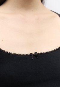 Close-up van het sleutelbeen van een persoon die een zwart topje draagt met kanten rand en een klein decoratief zwart strikje bij de halslijn.