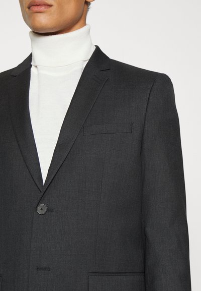 Dunkelgrauer Blazer aus strukturiertem Stoff, mit einem einzelnen Knopfverschluss und einer kleinen Brusttasche, getragen über einem weißen Rollkragenpullover.