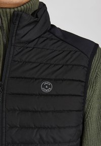 Sort polstret vest med lynlås, der har et rundt gråt logo. Skulderområdet er lavet af glat sort stof, og kraven er i olivengrønt tekstureret strik.