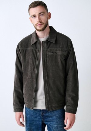 Brown corduroy jacket with a collared neckline, front zipper, and chest pocket, worn over a light-colored shirt and blue jeans.