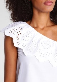 White sleeveless top featuring a large scalloped eyelet lace ruffle along the neckline, with soft cotton texture and fitted silhouette.