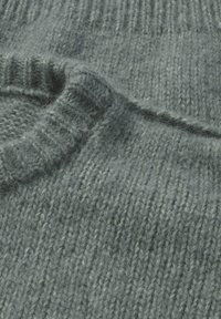 Grüner, gestrickter Pulloverstoff mit einem strukturierten Rippmuster. Das Material wirkt weich und flauschig und zeigt subtile Farbvariationen.