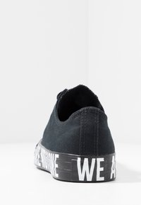 Chaussure en toile noire à profil bas, surface texturée et semelle extérieure en caoutchouc avec l'inscription "WE ARE" en lettres blanches épaisses.