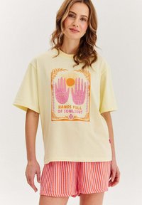 Žluté bavlněné tričko s grafikou růžových rukou a slunce, s nápisem "RUKY PLNÉ SLUNEČNÍHO SVĚTA", doplněné proužkovanými růžovými a oranžovými šortkami.