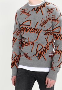 Persona che indossa una felpa grigia con grandi scritte arancioni e nere "Superdry", abbinata a jeans neri e una maglietta bianca visibile.